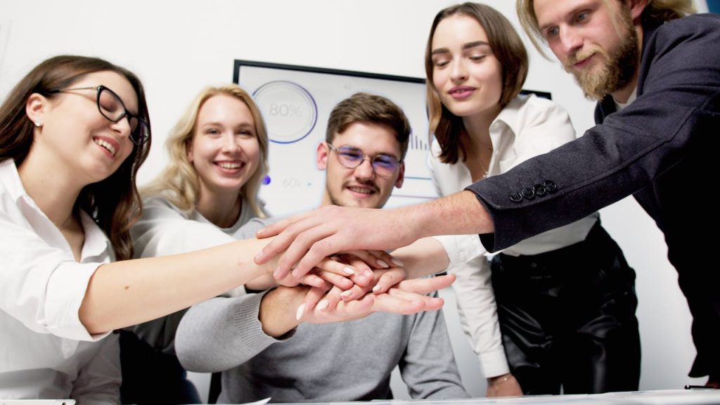 a-group-of-people-huddle-during-a-meeting-5716028 A diverse group of colleagues stacking hands in a gesture of teamwork and unity in an office setting.